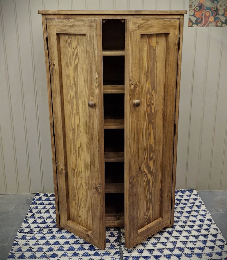 May include: A tall, rectangular wooden cupboard with a medium brown stained finish and visible wood grain. It features two hinged doors with small round knobs, opening to reveal five internal shelves stacked vertically inside the unit.