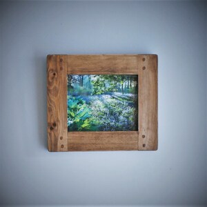 May include: A square wooden picture frame displaying a forest landscape photograph. The frame is constructed from light brown wood, showcasing the wood grain. The photograph depicts green leaves and a field of bluebell flowers.