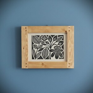 May include: A framed black and white floral print. The artwork features stylized flowers and leaves. The print is set in a natural wood frame, mounted on a blue wall. The frame is square and has visible wood grain.