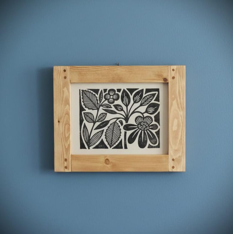 May include: A framed black and white floral print. The artwork features stylized flowers and leaves. The print is set in a natural wood frame, mounted on a blue wall. The frame is square and has visible wood grain.