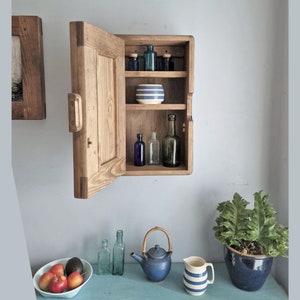 May include: A wooden cabinet with a door open, revealing three shelves with various glass bottles and a striped bowl. The cabinet is mounted on a wall.
