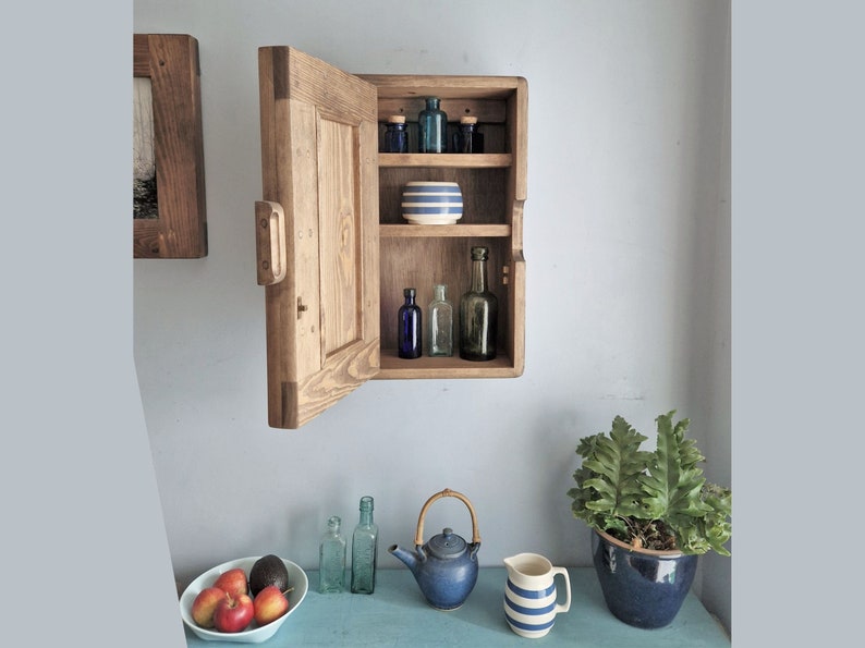 May include: A wooden cabinet with a door open, revealing three shelves with various glass bottles and a striped bowl. The cabinet is mounted on a wall.