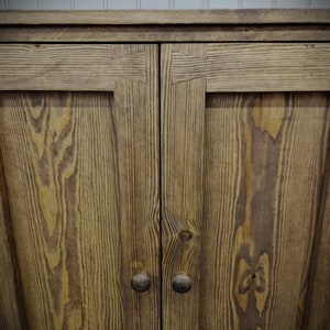 May include: A wooden cabinet featuring two doors with visible grain patterns and dark brown circular knobs. The upper frame of each door is constructed with prominent dovetail joinery, showcasing the natural texture of the wood.