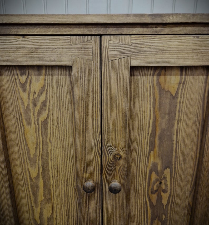 May include: A wooden cabinet featuring two doors with visible grain patterns and dark brown circular knobs. The upper frame of each door is constructed with prominent dovetail joinery, showcasing the natural texture of the wood.