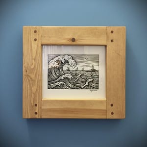 May include: A framed black and white print of ocean waves, sailboats, and a lighthouse. The artwork is in a light-colored wooden frame with visible wood grain and corner joinery. The print depicts a seascape with a large breaking wave.