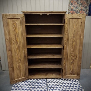 May include: A tall, rectangular wooden cabinet with a medium brown stained finish and visible wood grain. The cabinet features two hinged doors that are open, revealing five interior shelves for storage. The structure is made of solid wood planks.