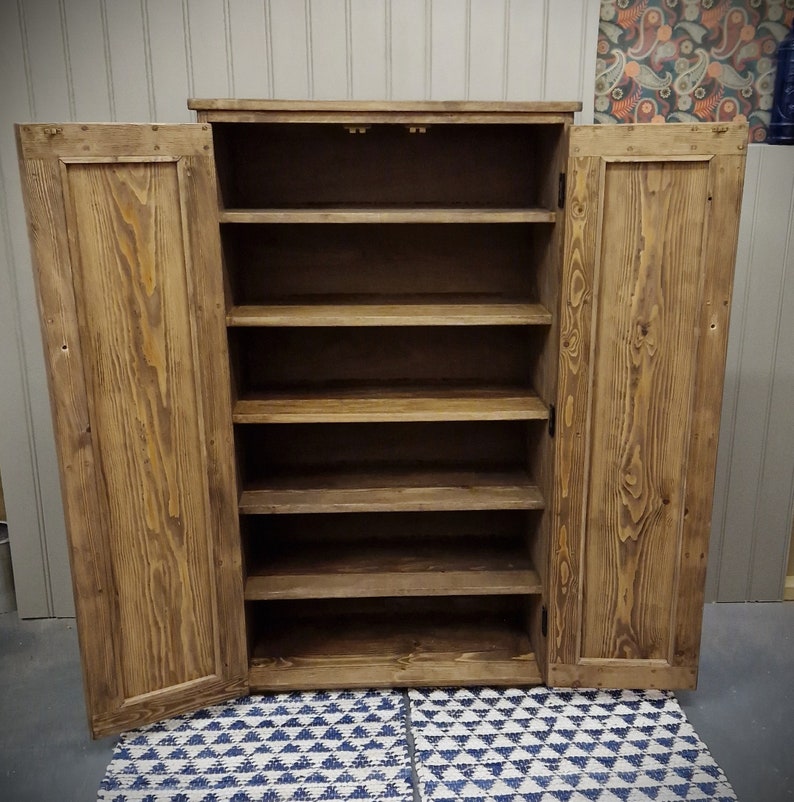 May include: A tall, rectangular wooden cabinet with a medium brown stained finish and visible wood grain. The cabinet features two hinged doors that are open, revealing five interior shelves for storage. The structure is made of solid wood planks.