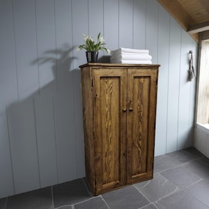 May include: A tall, rectangular wooden cabinet with two doors featuring a visible brown wood grain finish and small round knobs. A black pot containing white flowers and a stack of folded white towels sit on the flat top surface.