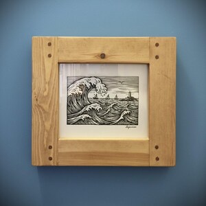 May include: A framed black and white print depicting a seascape with a large wave, sailboats, and a lighthouse. The artwork is in a light-coloured wooden frame with visible wood grain and corner joinery. The print is a detailed illustration.