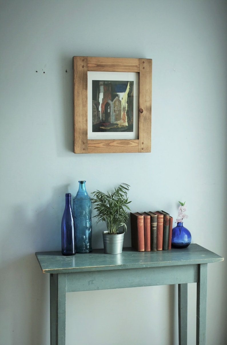 May include: A wooden framed print of a building hangs above a teal table. On the table are two blue glass bottles, a potted plant, several books, and a small blue vase with flowers. The scene is set against a light blue wall.
