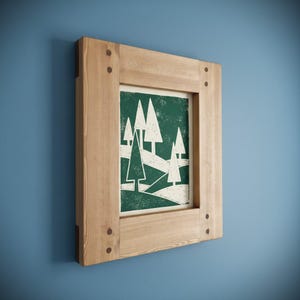 May include: A framed art print featuring a stylized forest scene with white trees on a dark green background. The artwork is encased in a natural wood frame, secured with visible pegs at the corners. The frame is mounted on a blue wall.