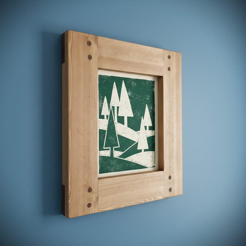 May include: A framed art print featuring a stylized forest scene with white trees on a dark green background. The artwork is encased in a natural wood frame, secured with visible pegs at the corners. The frame is mounted on a blue wall.