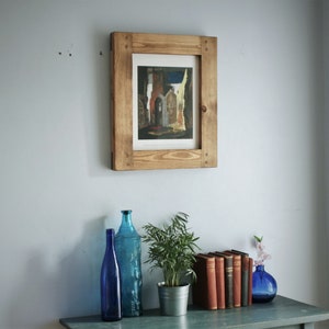 May include: A framed print of a building with a wooden frame is mounted on a light blue wall. Below, a blue table holds blue glass bottles, a potted plant in a metal pot, a stack of books, and a blue vase with flowers.