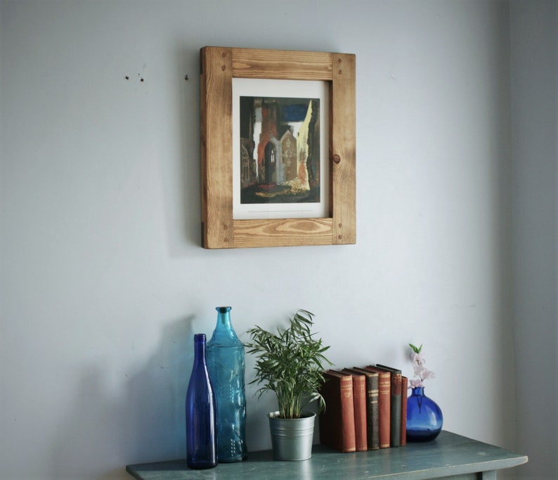 May include: A framed print of a building with a wooden frame is mounted on a light blue wall. Below, a blue table holds blue glass bottles, a potted plant in a metal pot, a stack of books, and a blue vase with flowers.