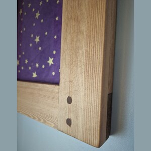 May include: Wooden framed artwork featuring a purple background with gold stars. The frame is made of light-colored wood with visible grain and is secured with two dark metal pins. The artwork is mounted on a light blue wall.
