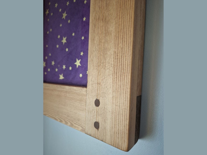 May include: Wooden framed artwork featuring a purple background with gold stars. The frame is made of light-colored wood with visible grain and is secured with two dark metal pins. The artwork is mounted on a light blue wall.