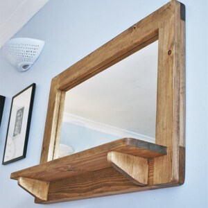 May include: A rustic wooden mirror with a shelf underneath. The mirror has a rectangular frame and the shelf is made of the same wood. The wood is a dark brown color and has a natural finish.