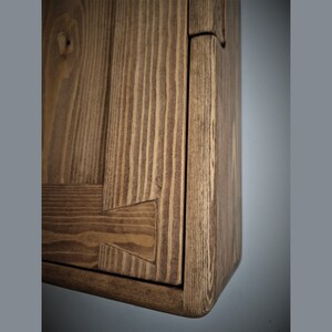 Puede incluir: Primer plano de una esquina de un armario de madera con un acabado marr&oacute;n natural. El armario tiene un dise&ntilde;o sencillo con un borde biselado y un patr&oacute;n de veta visible.