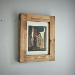 May include: A framed print of a building with a brown, light brown, and red color scheme. The frame is made of light brown wood and is mounted on a gray wall. The print is rectangular and has a white border.