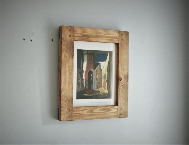May include: A framed print of a building with a brown, light brown, and red color scheme. The frame is made of light brown wood and is mounted on a gray wall. The print is rectangular and has a white border.
