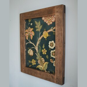 May include: A framed wall art piece featuring a dark teal fabric with a floral pattern in shades of yellow, pink, and green. The fabric is set within a rustic, stained wooden frame, creating a decorative accent for home decor.