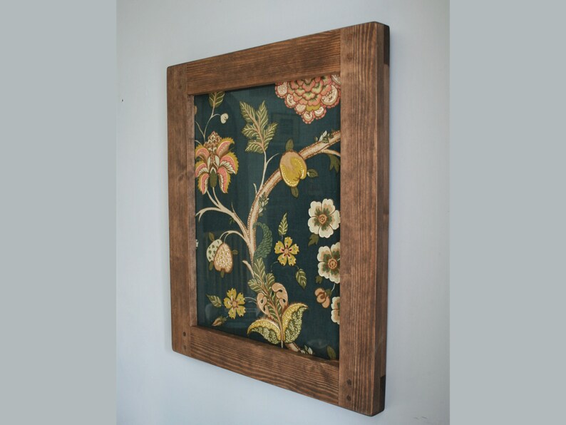 May include: A framed wall art piece featuring a dark teal fabric with a floral pattern in shades of yellow, pink, and green. The fabric is set within a rustic, stained wooden frame, creating a decorative accent for home decor.