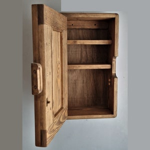 May include: A wooden wall-mounted cabinet with a single door and two shelves. The door is open, revealing the empty shelves inside. The cabinet is made of unfinished wood with a natural brown color.