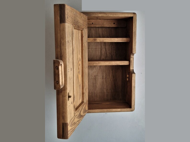 May include: A wooden wall-mounted cabinet with a single door and two shelves. The door is open, revealing the empty shelves inside. The cabinet is made of unfinished wood with a natural brown color.