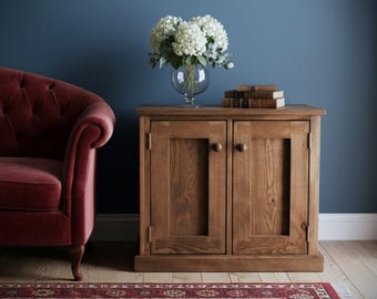 May include: A wooden cabinet with two doors and a natural finish. A glass vase with white flowers sits on top, along with a stack of books. A red armchair and patterned rug are in the foreground, against a dark blue wall.