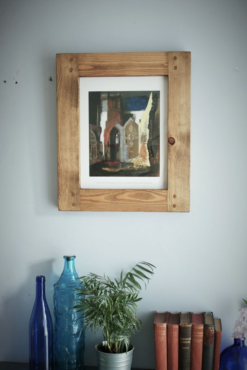May include: A framed print of a building with a light brown wooden frame. The print depicts a building with arched windows and a dark blue sky. The frame is made of light brown wood with visible wood grain.