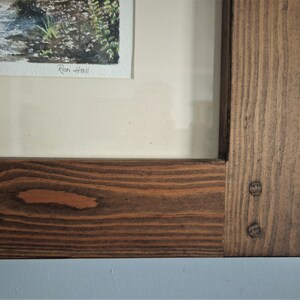 May include: A framed watercolor painting of a stream with a wooden frame. The frame is made of stained wood with visible wood grain and two decorative pegs. The artist's signature, "Ron Hall", is visible in the painting.