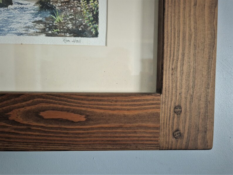 May include: A framed watercolor painting of a stream with a wooden frame. The frame is made of stained wood with visible wood grain and two decorative pegs. The artist's signature, "Ron Hall", is visible in the painting.