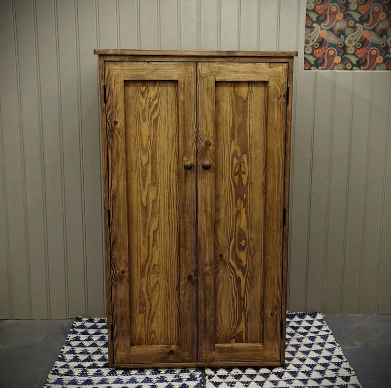 May include: A tall, rectangular wooden cabinet with a medium-brown stained finish and visible wood grain. The cabinet features two vertical doors, each equipped with a small, round wooden knob centred on the door panel.
