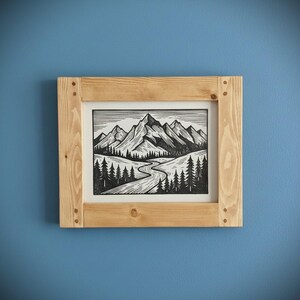 May include: A framed black and white mountain landscape art print. The artwork depicts a winding river, pine trees, and towering mountains. The frame is made of light-colored wood and is mounted on a blue wall.