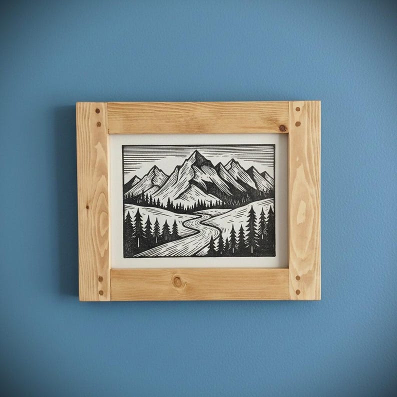 May include: A framed black and white mountain landscape art print. The artwork depicts a winding river, pine trees, and towering mountains. The frame is made of light-colored wood and is mounted on a blue wall.