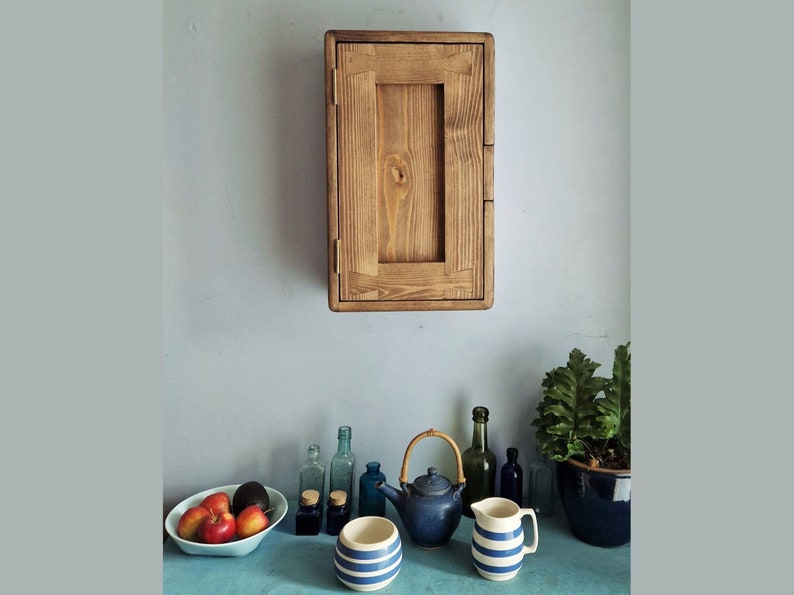 May include: A wooden wall-mounted cabinet with a single door. The cabinet is made of light brown wood and has a simple design. The cabinet is mounted on a light blue wall. There are several bottles, a teapot, and a pitcher on a blue countertop in front of the cabinet.