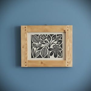 May include: A framed black and white floral print. The artwork features stylized flowers and leaves. The print is set within a natural wood frame, mounted on a blue wall. The frame is constructed with visible corner joints.