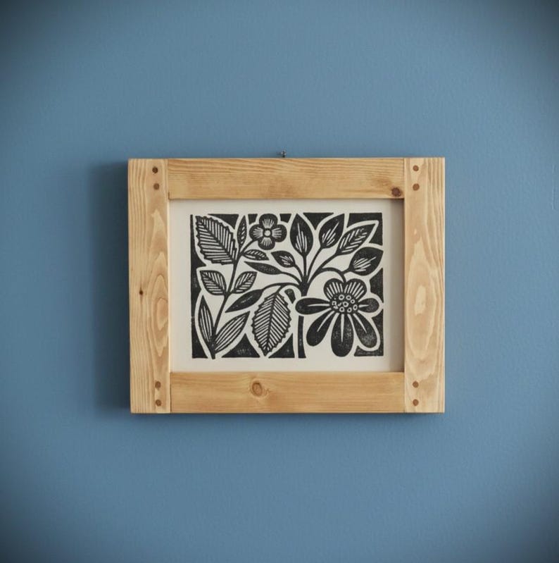 May include: A framed black and white floral print. The artwork features stylized flowers and leaves. The print is set within a natural wood frame, mounted on a blue wall. The frame is constructed with visible corner joints.