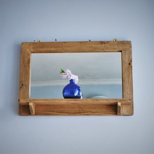 May include: A wooden shelf with a mirror and a blue vase with pink flowers. The shelf is mounted on a wall.
