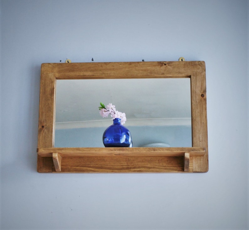 May include: A wooden shelf with a mirror and a blue vase with pink flowers. The shelf is mounted on a wall.