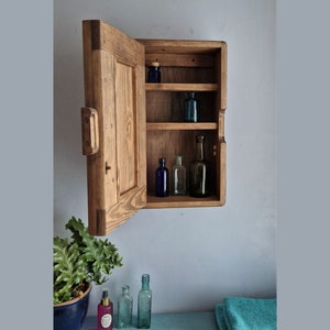 Puede incluir: Un botiqu&iacute;n de madera con una puerta con bisagras abierta, que revela tres estantes con botellas de vidrio. El botiqu&iacute;n est&aacute; montado en una pared y tiene un acabado r&uacute;stico y desgastado.