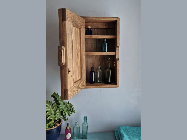 Puede incluir: Un botiqu&iacute;n de madera con una puerta con bisagras abierta, que revela tres estantes con botellas de vidrio. El botiqu&iacute;n est&aacute; montado en una pared y tiene un acabado r&uacute;stico y desgastado.