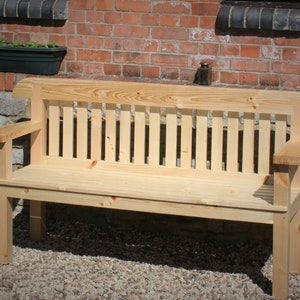 May include: A light-colored wooden bench with a slatted back and armrests. The bench is made of unfinished wood and is set against a brick wall. The bench is approximately 60 inches long.