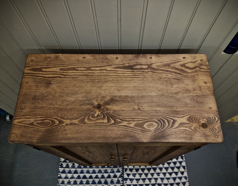 May include: A rectangular wooden cabinet top with a dark brown stained finish, showcasing prominent wood grain patterns, knots, and small circular metal nail heads embedded along the edges of the flat surface.