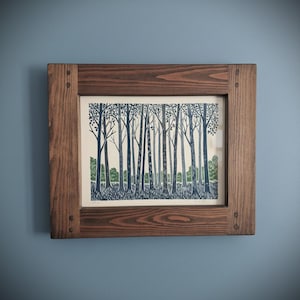 May include: A framed art print featuring a forest scene. The artwork depicts tall trees in shades of blue and green against a cream background. The print is encased in a rustic, dark brown wooden frame, mounted on a blue wall.