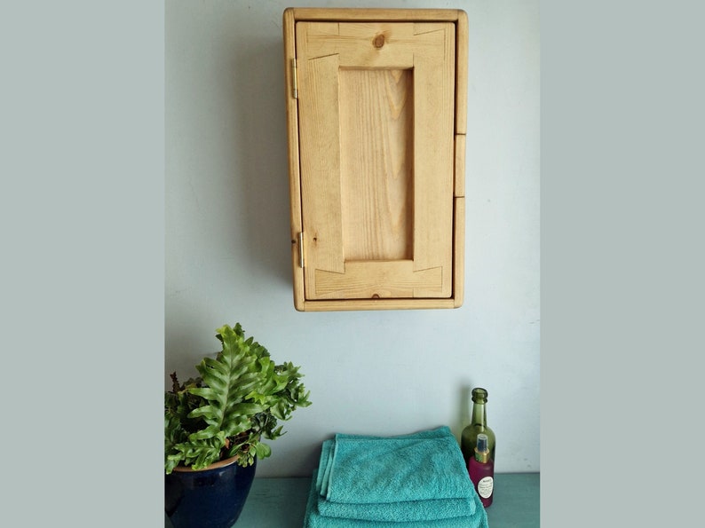 Puede incluir: Un armario de madera con una sola puerta, montado en una pared azul claro. El armario est&aacute; hecho de madera clara con un acabado natural. La puerta est&aacute; cerrada y tiene un dise&ntilde;o sencillo. Hay dos toallas y una botella de l&iacute;quido en un estante debajo del armario.