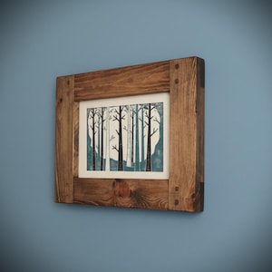 May include: A framed art print depicting a forest scene with black trees on a teal backdrop. The print is set within a rustic, square wooden frame, showing wood grain and corner joints. The artwork is mounted on a blue wall.