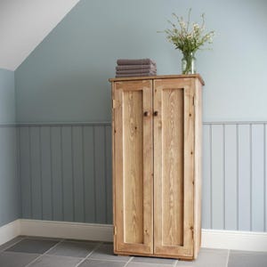 May include: A tall, light-toned wooden cabinet with two vertical doors, each featuring a single round knob. A stack of folded grey towels and a clear glass vase containing a bouquet of white wildflowers sit on the top surface.