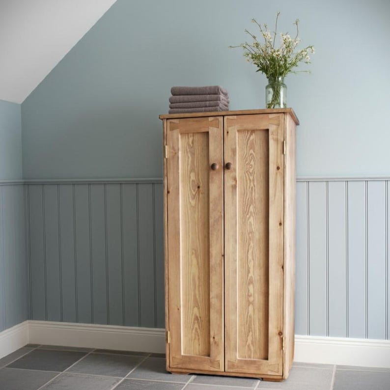 May include: A tall, light-toned wooden cabinet with two vertical doors, each featuring a single round knob. A stack of folded grey towels and a clear glass vase containing a bouquet of white wildflowers sit on the top surface.