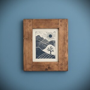 May include: A framed print depicting a landscape with hills, a tree, and a sun. The artwork uses blue and white tones, encased in a rectangular wooden frame. The frame is made of natural wood and is mounted on a blue wall.